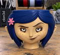 Coraline candle holder with dark blue hair, a pink flower accent, and a freckled face on a workbench.
