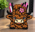 Highland cow pencil holder with brown shaggy detailing, black horns, and a pink flower, holding pens on a desk.