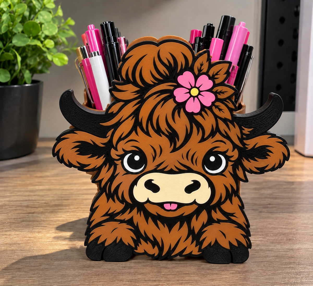 Highland cow pencil holder with brown shaggy detailing, black horns, and a pink flower, holding pens on a desk.