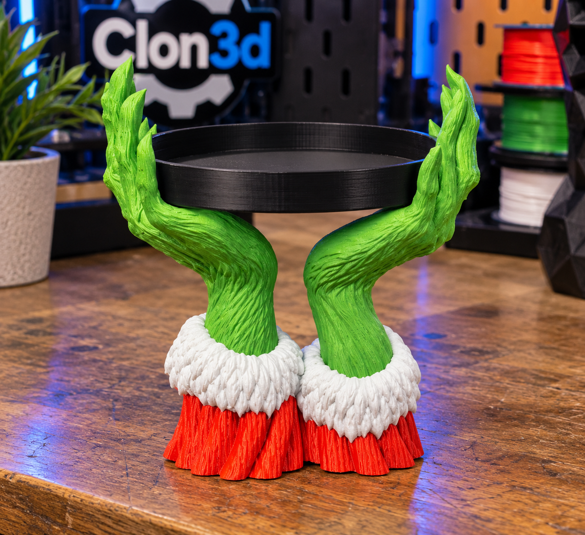 Grinch hands candle holder with green hands, red sleeves, white cuffs, and a black circular tray on a workbench.